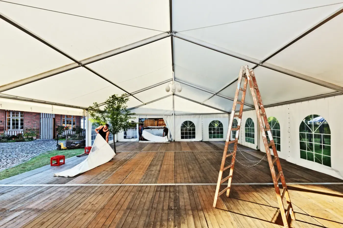Eine Person baut ein großes weißes Festzelt mit Holzboden, Holzleiter und Bogenfenstern auf.