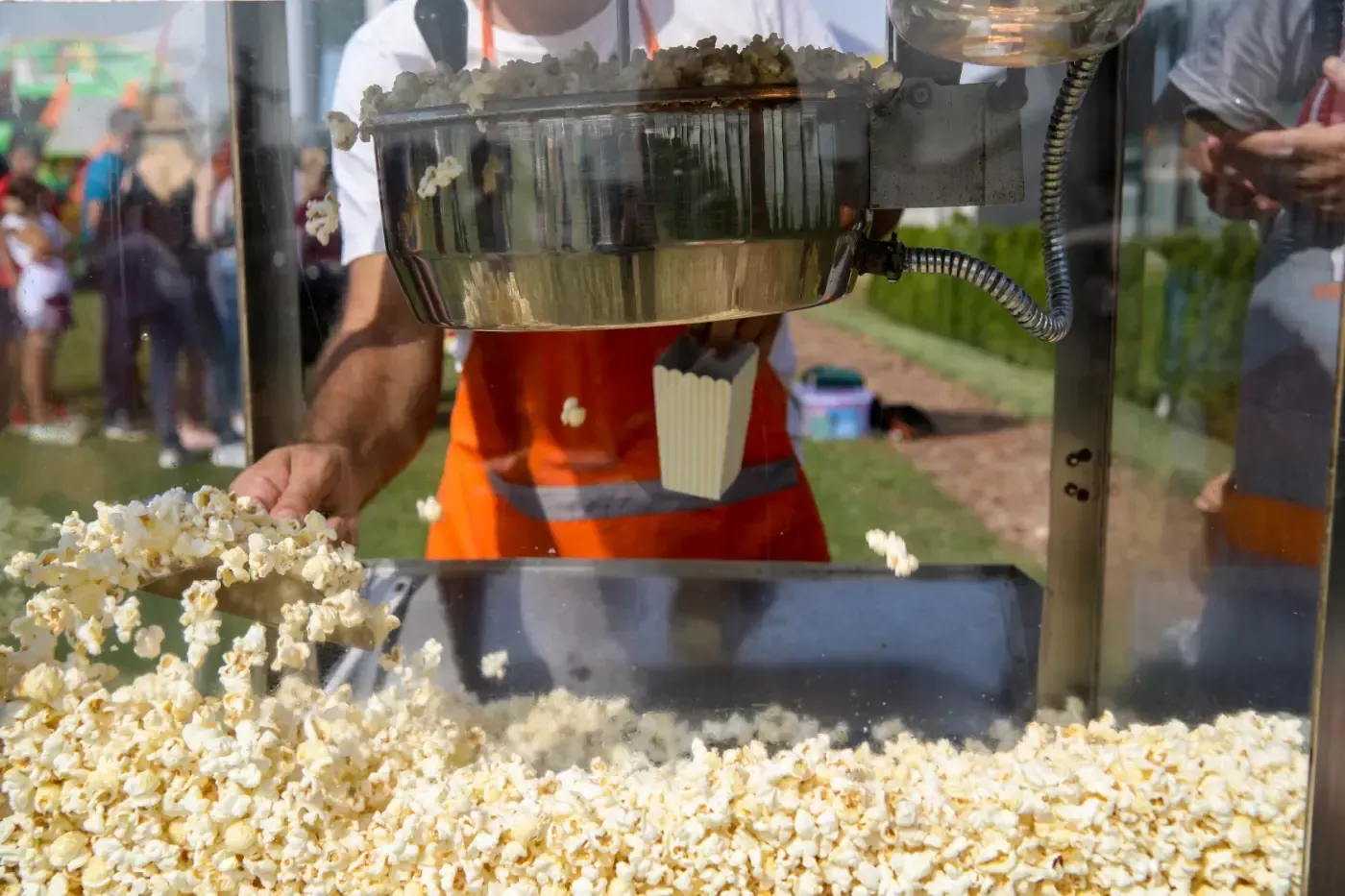 Eine Person in einer orangefarbenen Schürze serviert bei einer Freiluftveranstaltung mit einer Metallschaufel frisches Popcorn aus einer gewerblichen Maschine.