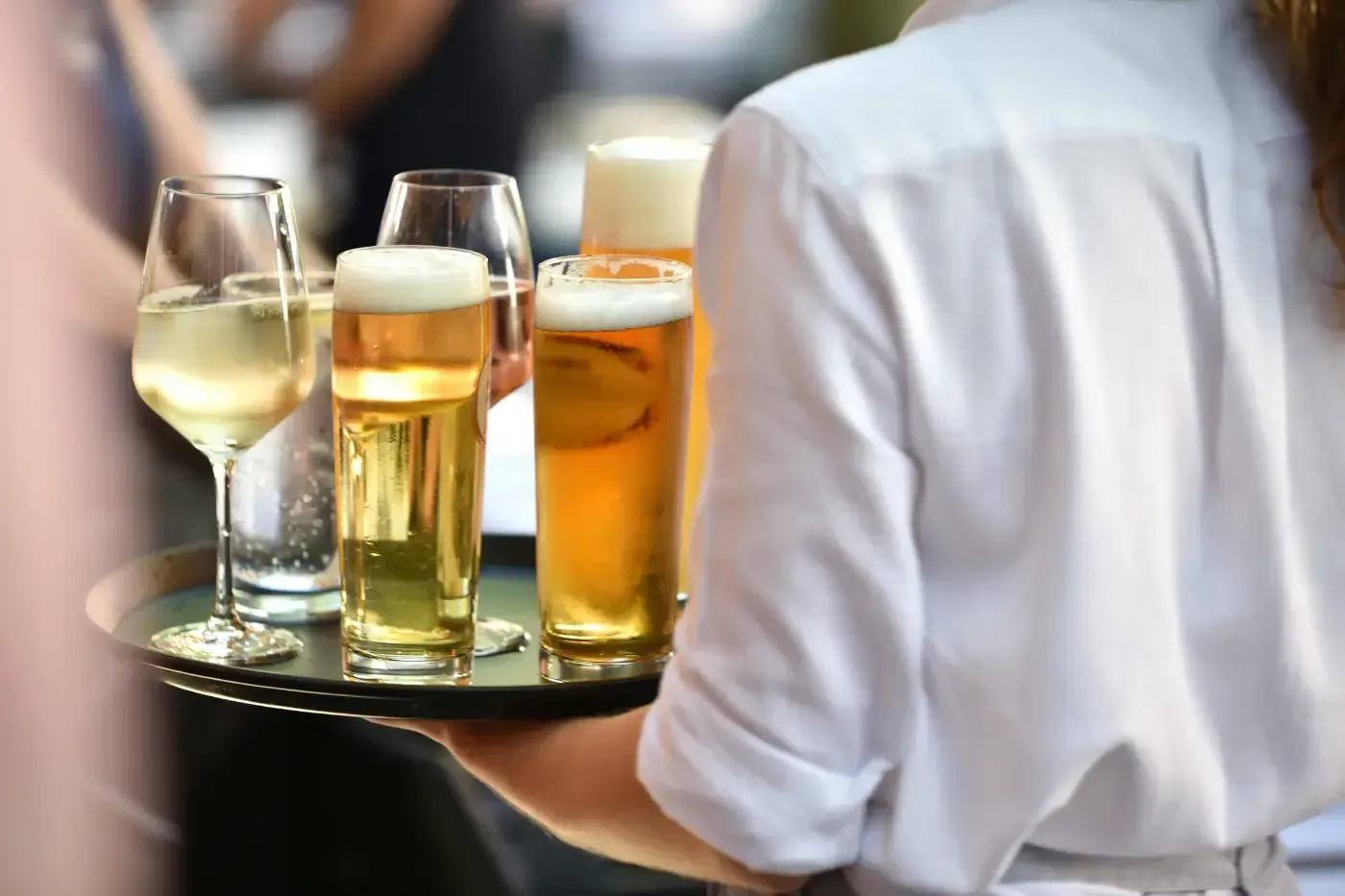 Ein Kellner in einem weißen Hemd trägt in einer Außenkulisse ein schwarzes Tablett mit Gläsern Bier und Weißwein.