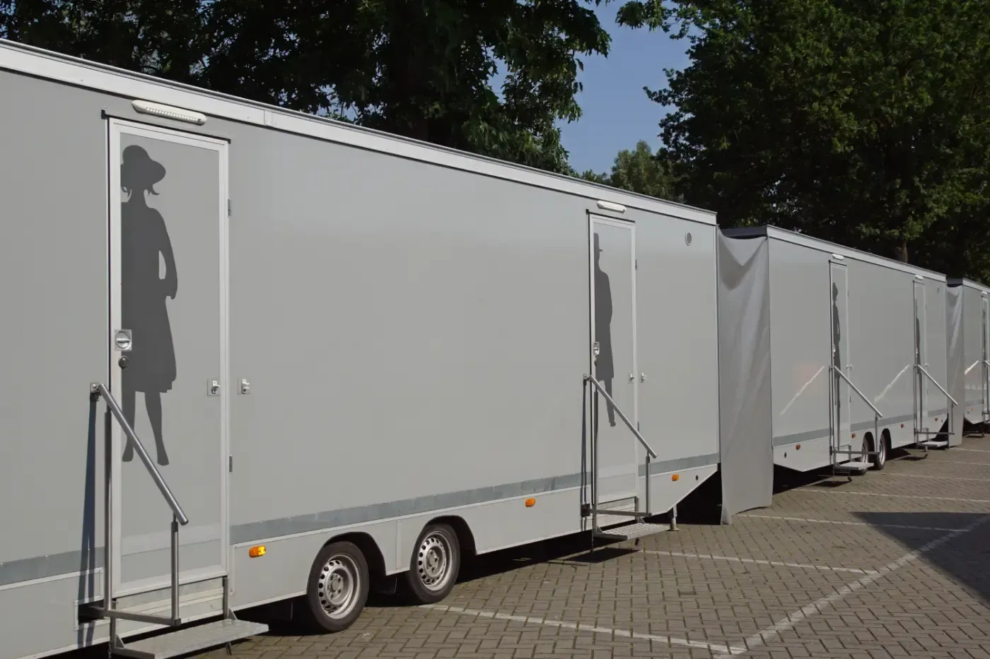 Eine Reihe hellgrauer, fahrbarer Toilettenwagen stand auf einem gepflasterten Parkplatz; die Türschilder zeigten Silhouette-Symbole.
