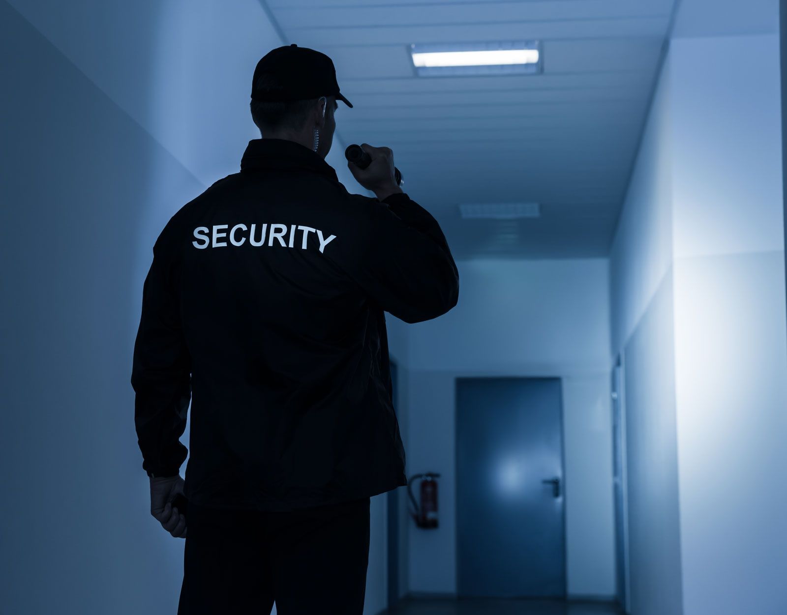 Ein Wachmann in dunkler Uniform mit einer Taschenlampe in der Hand steht in einem schwach beleuchteten, bläulich getönten Flur.