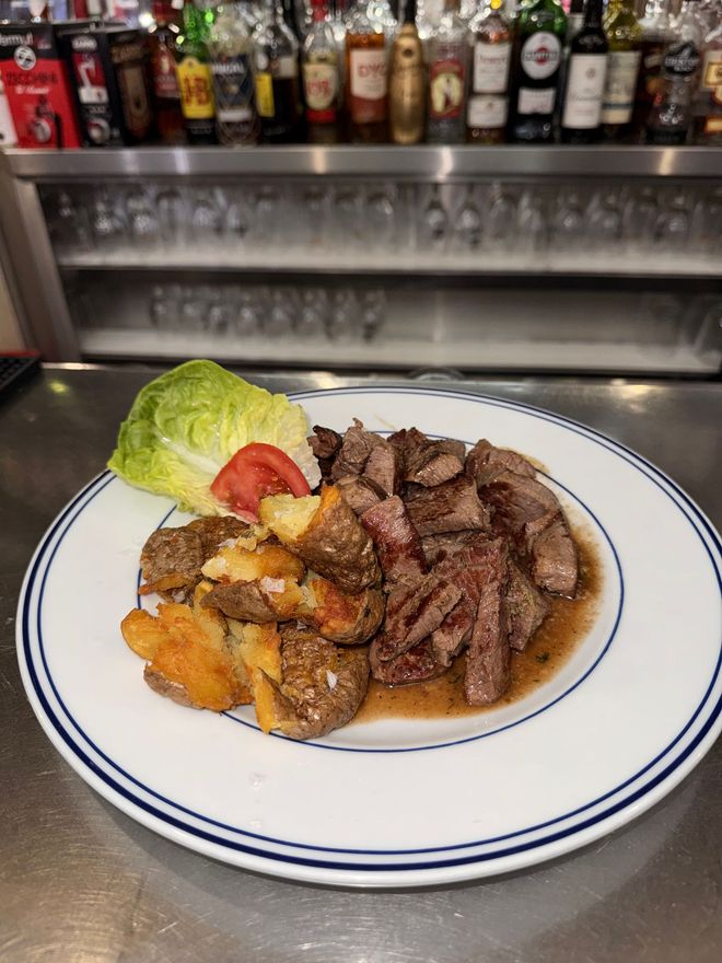 Filete emplatado con patatas asadas y guarnición de lechuga sobre la barra de un bar, con botellas de licor al fondo.