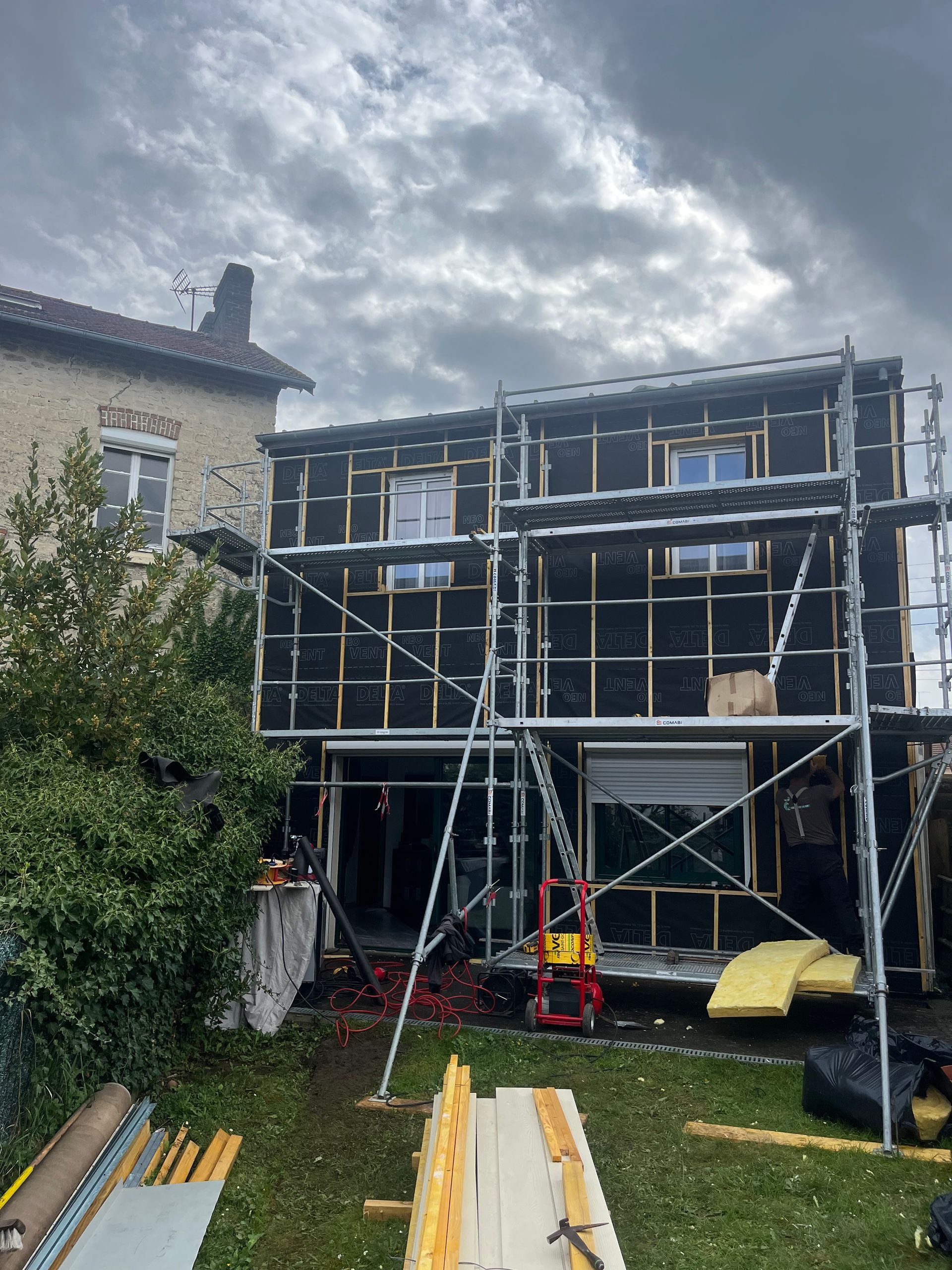 Échafaudage devant une petite maison côté jardin dont la façade est recouvert d'une bâche noir