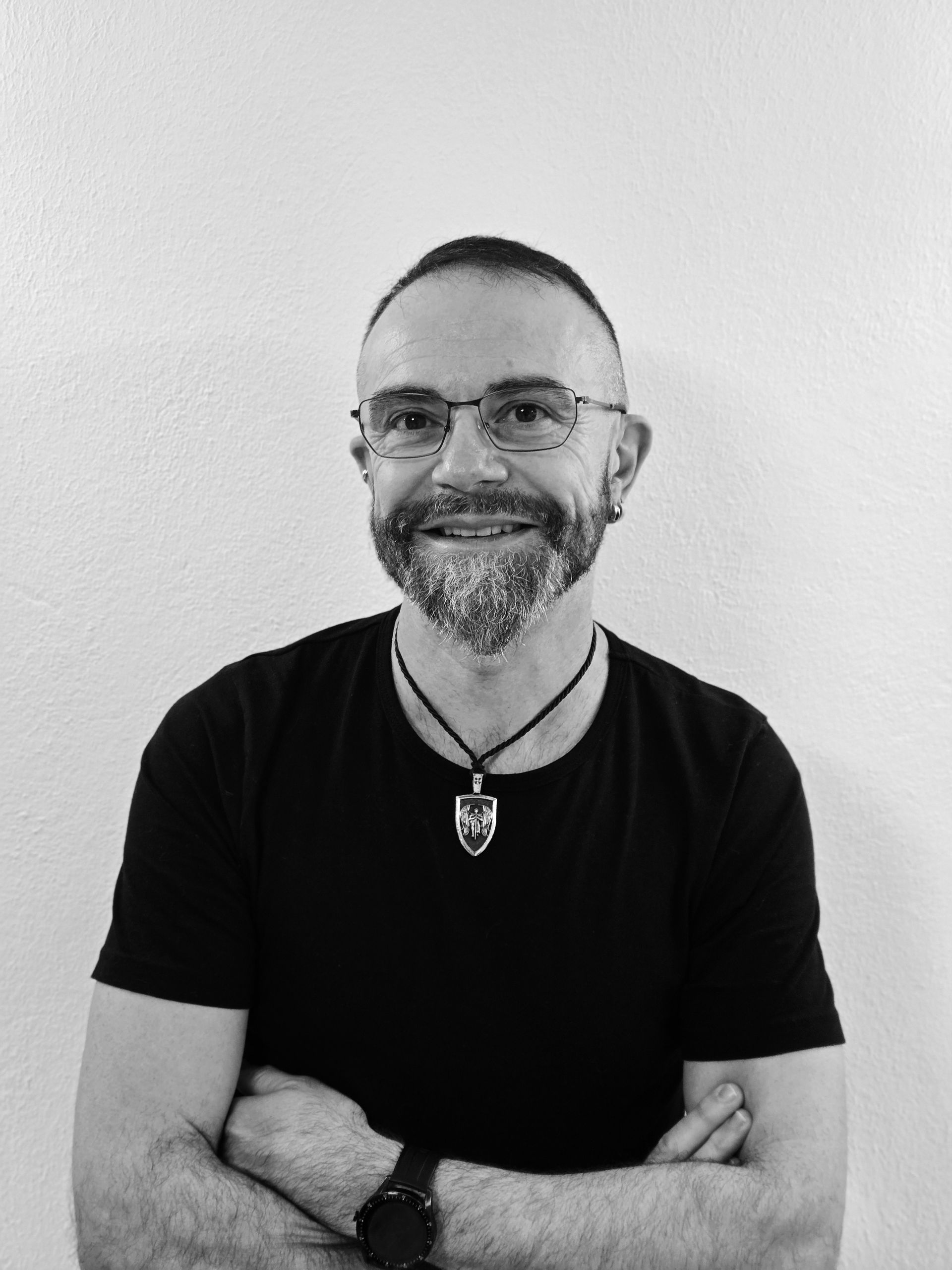 Mann mit Brille und Bart, die Arme verschränkt, lächelt. Er trägt ein schwarzes Hemd und einen Anhänger und steht vor einer weißen Wand.