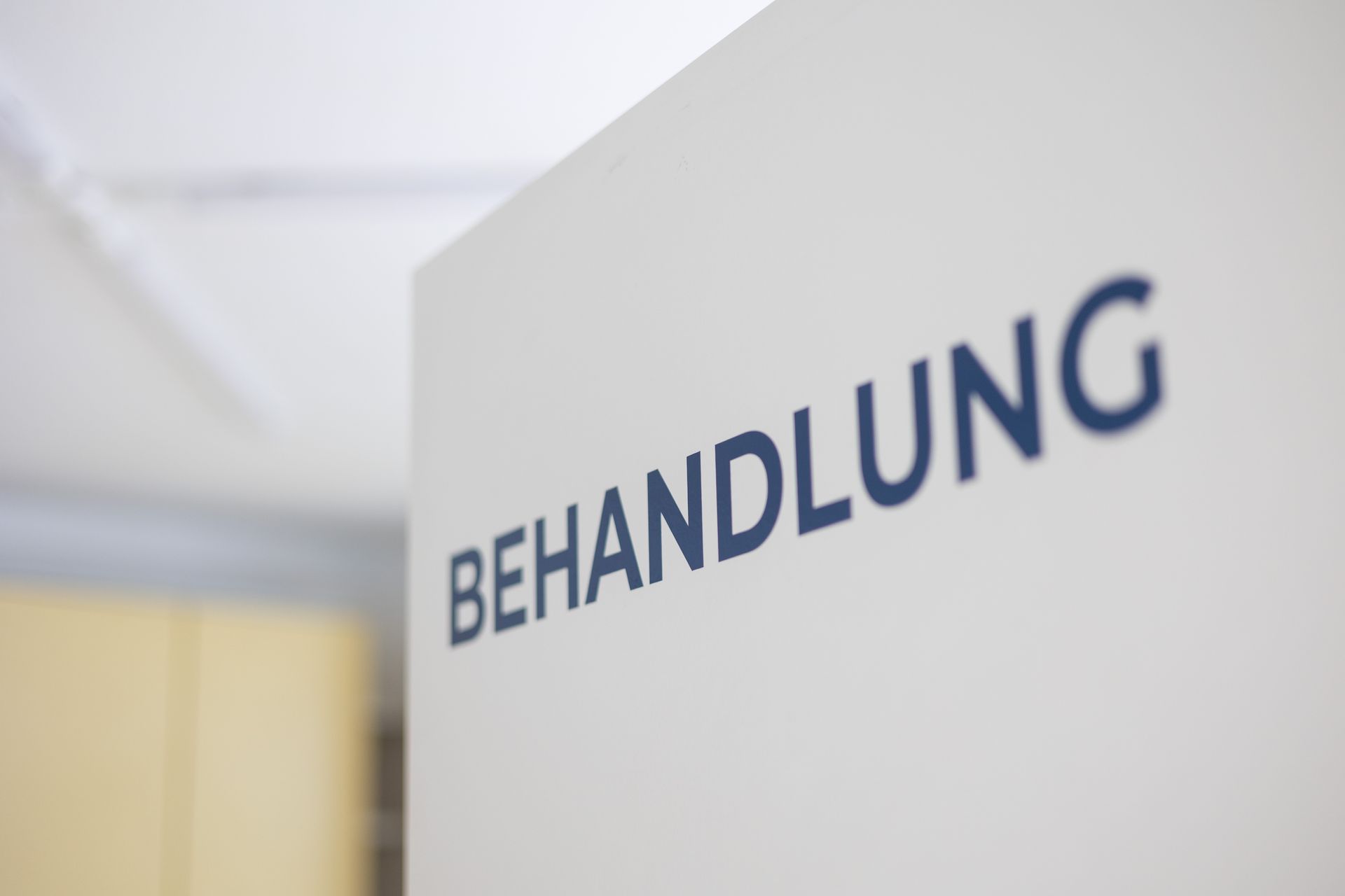 Weiße Wand mit dem Wort „BEHANDLUNG“ in dunkelblauen Buchstaben.