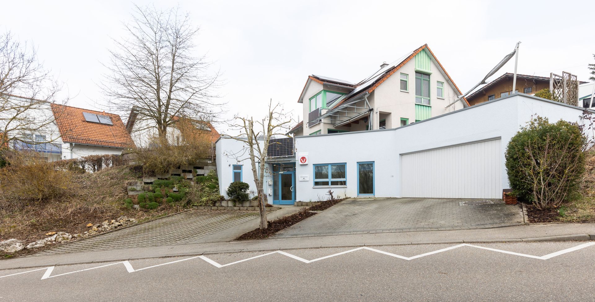 Weißes Gebäude mit blauer Tür, Fenstern und rotem Logo, im Hintergrund ein zweistöckiges Haus.