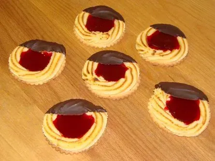 Sechs kleine, säuerliche Kekse mit roter Geleefüllung und dunkler Schokoladenhaube auf einem Holzsockel.