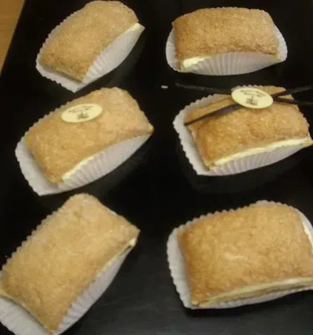 Sechs rechteckige, mit Kokosraspeln überzogene Gebäckstücke in weißen Papierförmchen auf dunklem Untergrund.