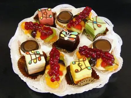 Verschiedene dekorierte Petit Fours auf einer weißen Deckchenplatte, garniert mit Früchten und Schokoladenglasur.
