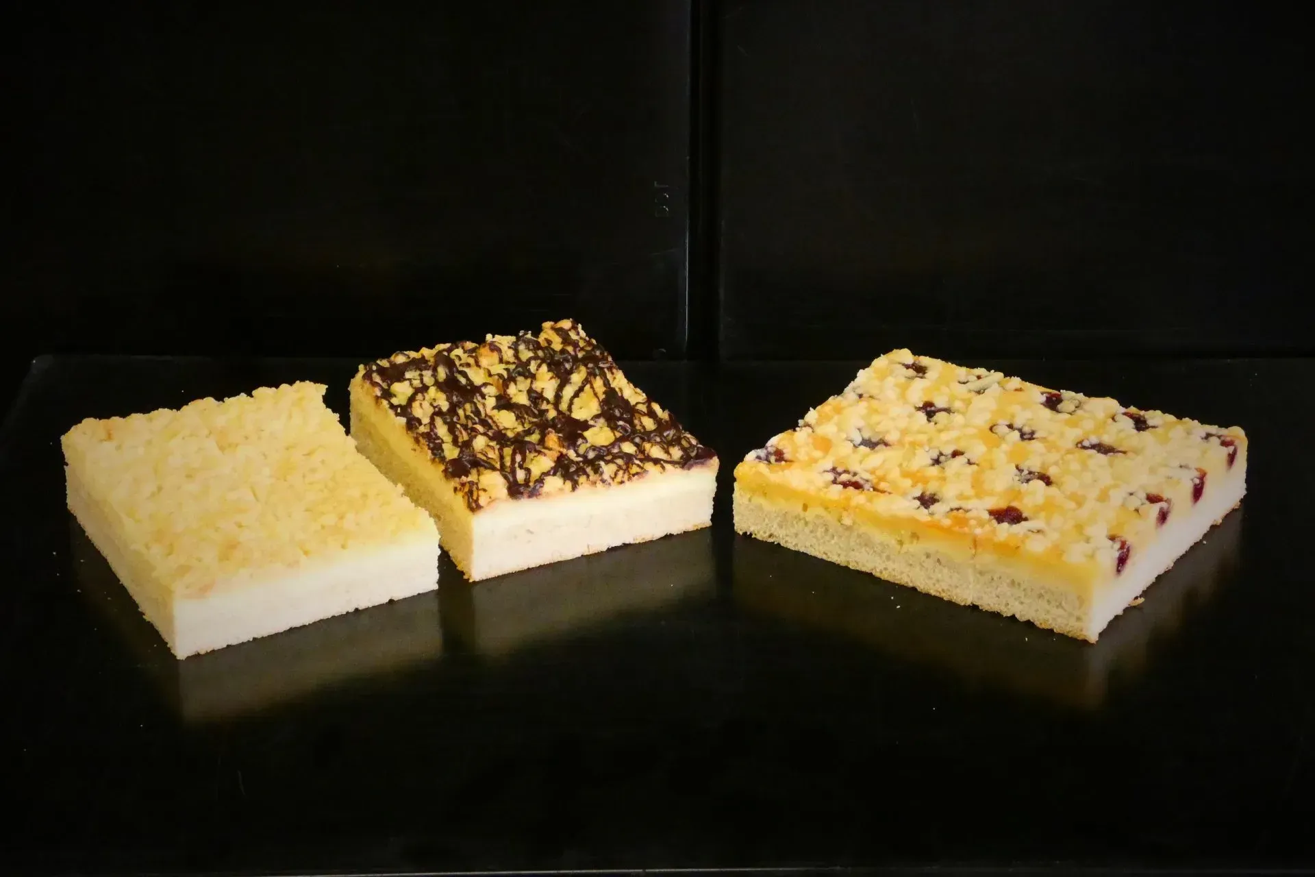 Drei quadratische Dessertriegel mit leichter Glasur und verschiedenen Toppings auf dunklem Untergrund