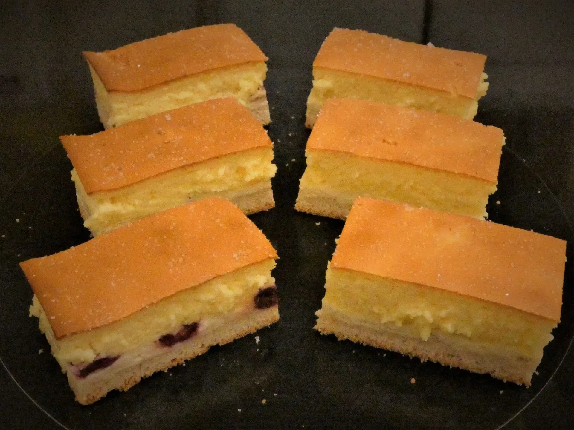 Sechs Scheiben Biskuitkuchen mit Orangenglasur auf einer dunklen Oberfläche angerichtet.