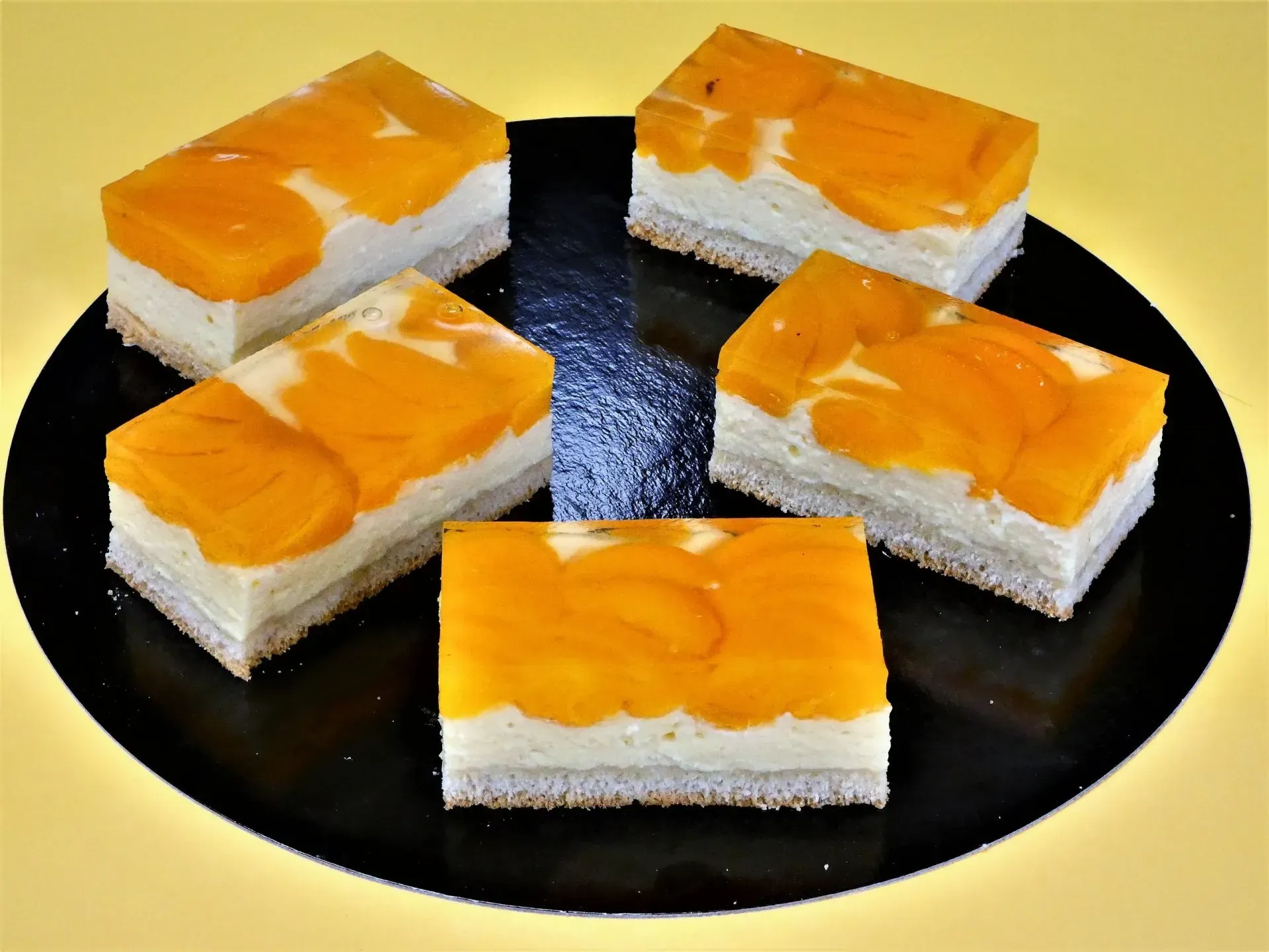 Fünf mit Früchten belegte Dessertquadrate auf einem schwarzen Teller vor gelbem Hintergrund