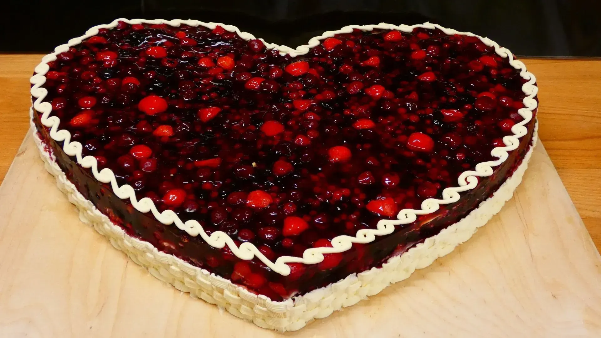 Herzförmige Beerentarte mit glänzenden roten Beeren und weißer Sahne auf einem Holzbrett