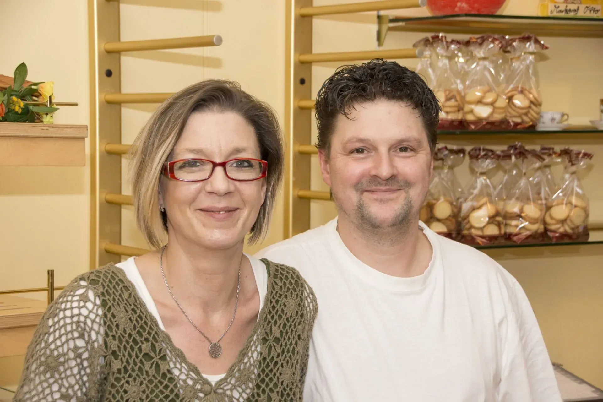Zwei lächelnde Personen in einer Bäckerei, im Hintergrund Regale voller Gebäck.