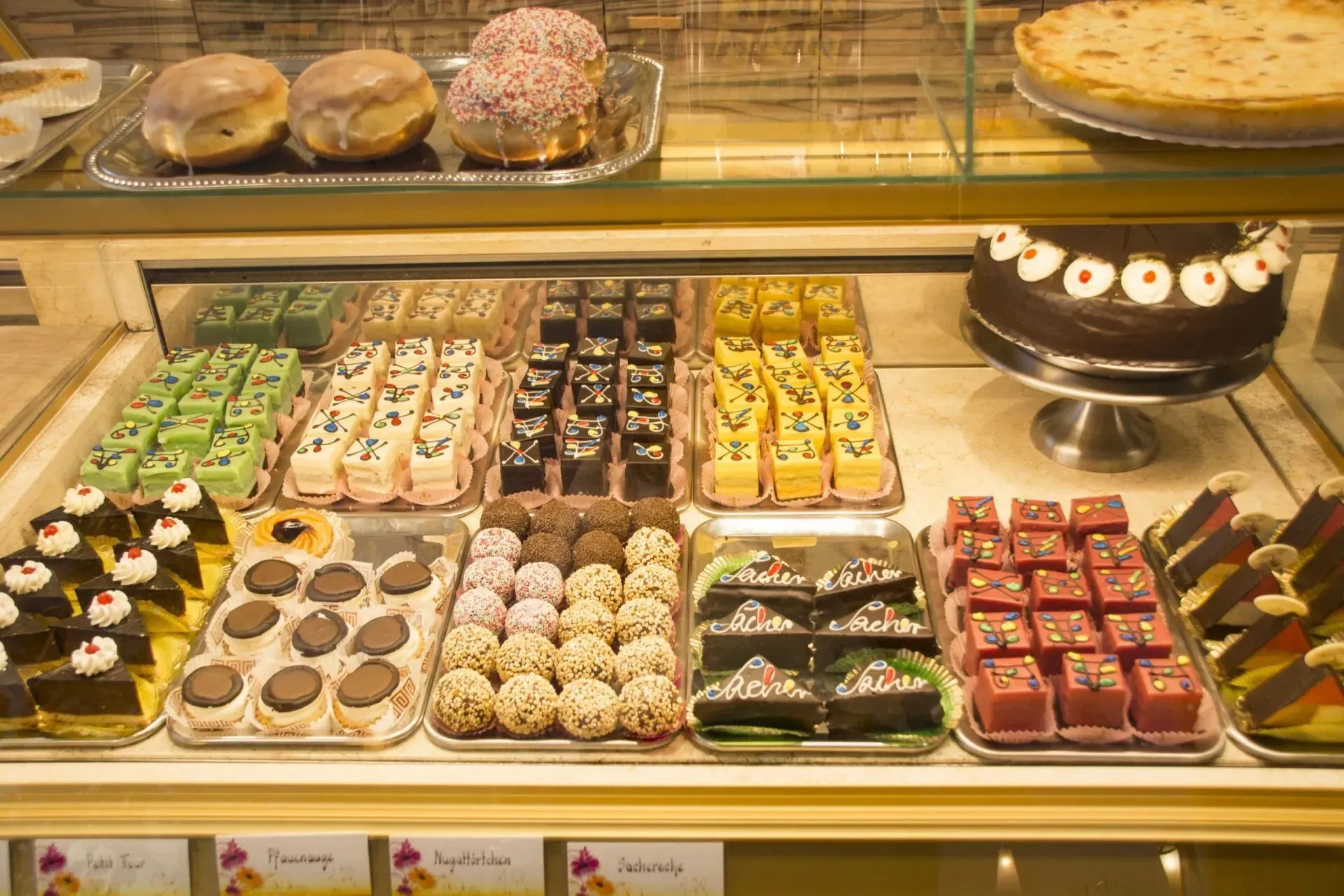 Auslage einer Bäckerei mit verschiedenen bunten Gebäckstücken, Kuchen und Törtchen, die in Reihen angeordnet sind.