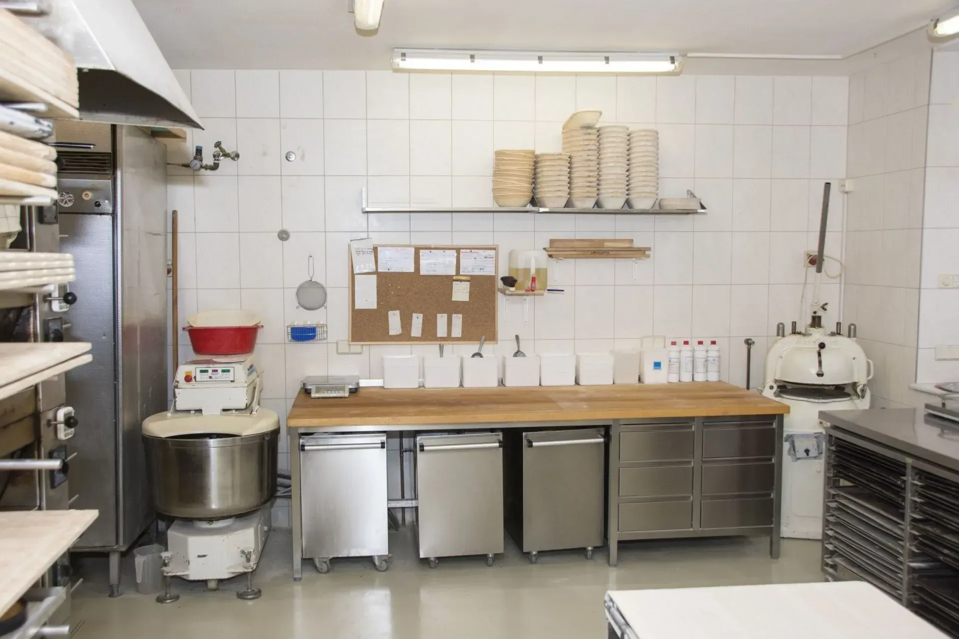 Gewerbliche Küche mit Arbeitsflächen, Mixern, Regalen und Aufbewahrungsbehältern aus Edelstahl an einer gefliesten Wand.