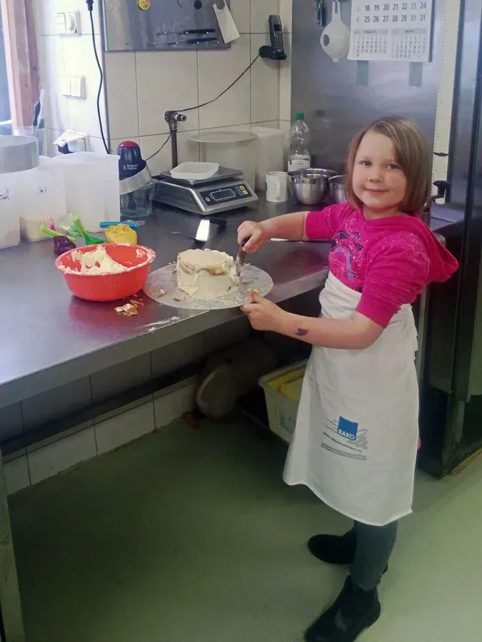 Ein Kind in einer weißen Schürze lächelt und hält eine Schüssel mit Kuchenmischung in der Hand in einer Küchenwerkstatt.
