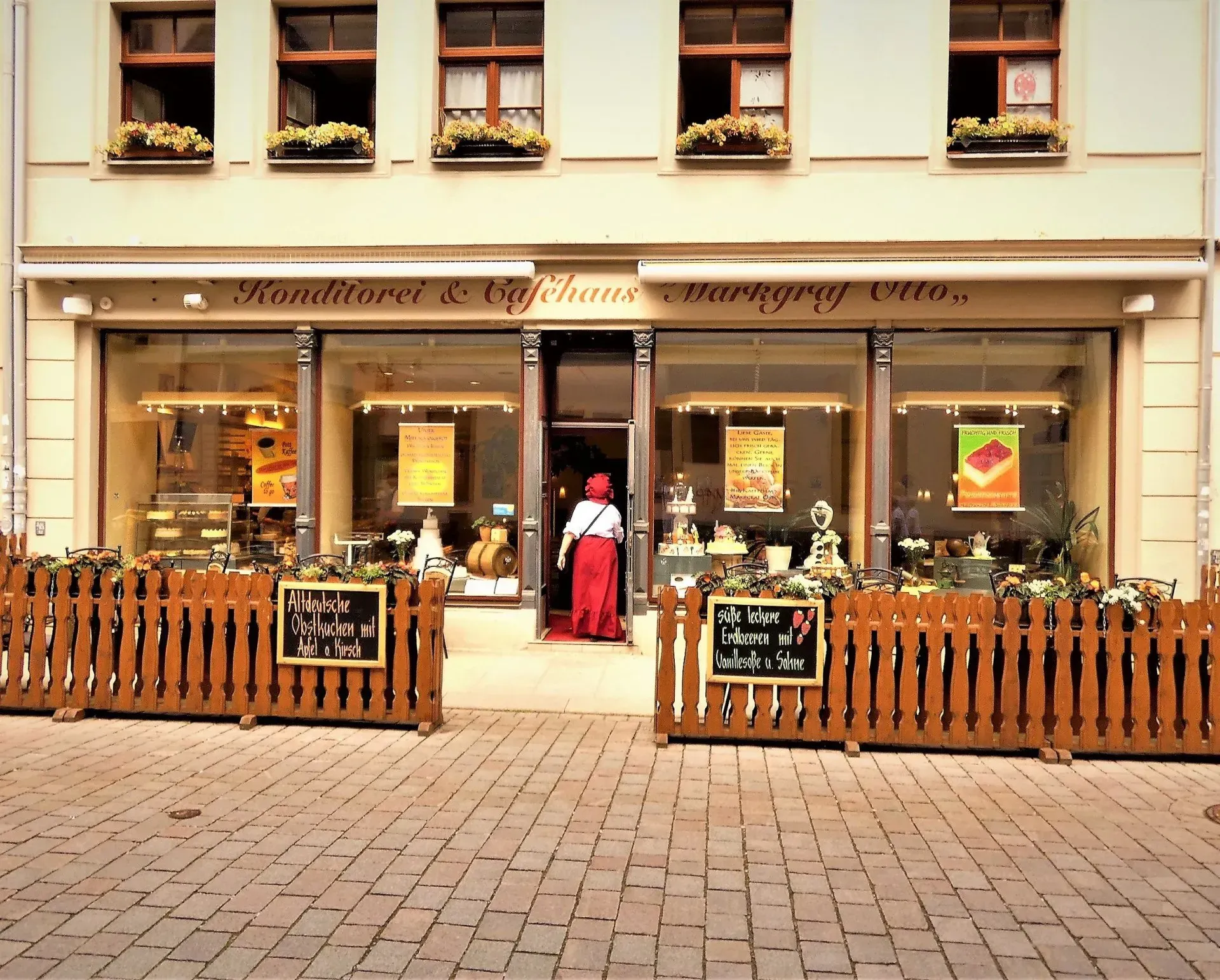Bäckerei oder Café mit Straßenzugang, Außensitzplätzen, Blumenkästen und Leuten, die am Eingang stehen.