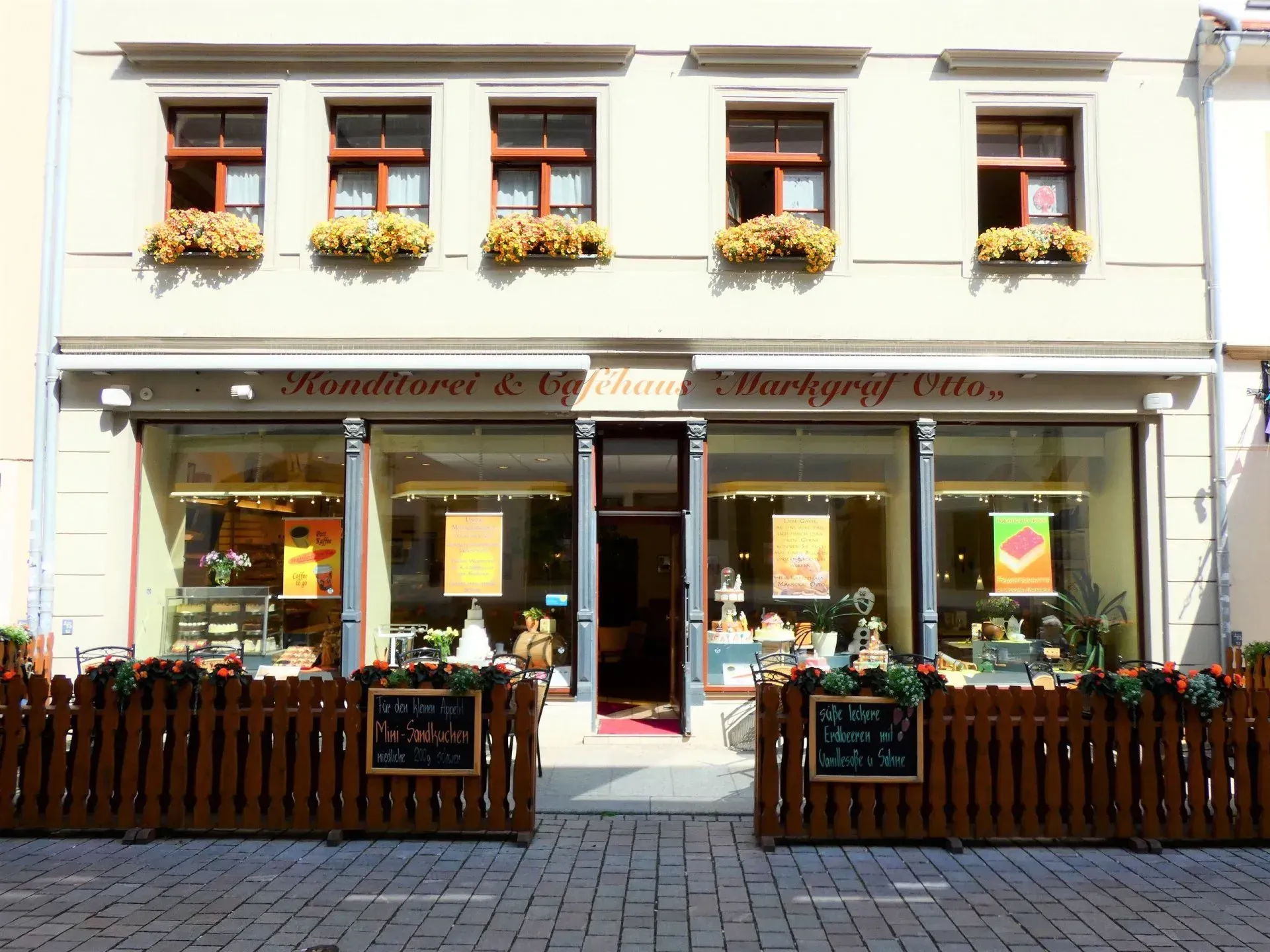 Vorderansicht eines malerischen Cafés mit Blumenkästen, großen Fenstern sowie einem hölzernen Außenzaun und Tischen.