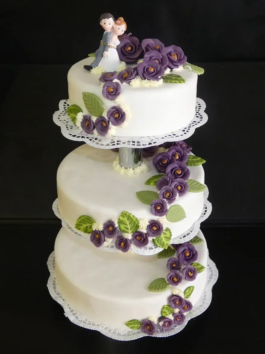 Dreistöckige weiße Hochzeitstorte mit lila Blüten und Spitzenbesatz, gekrönt von einer Brautpaarfigur.