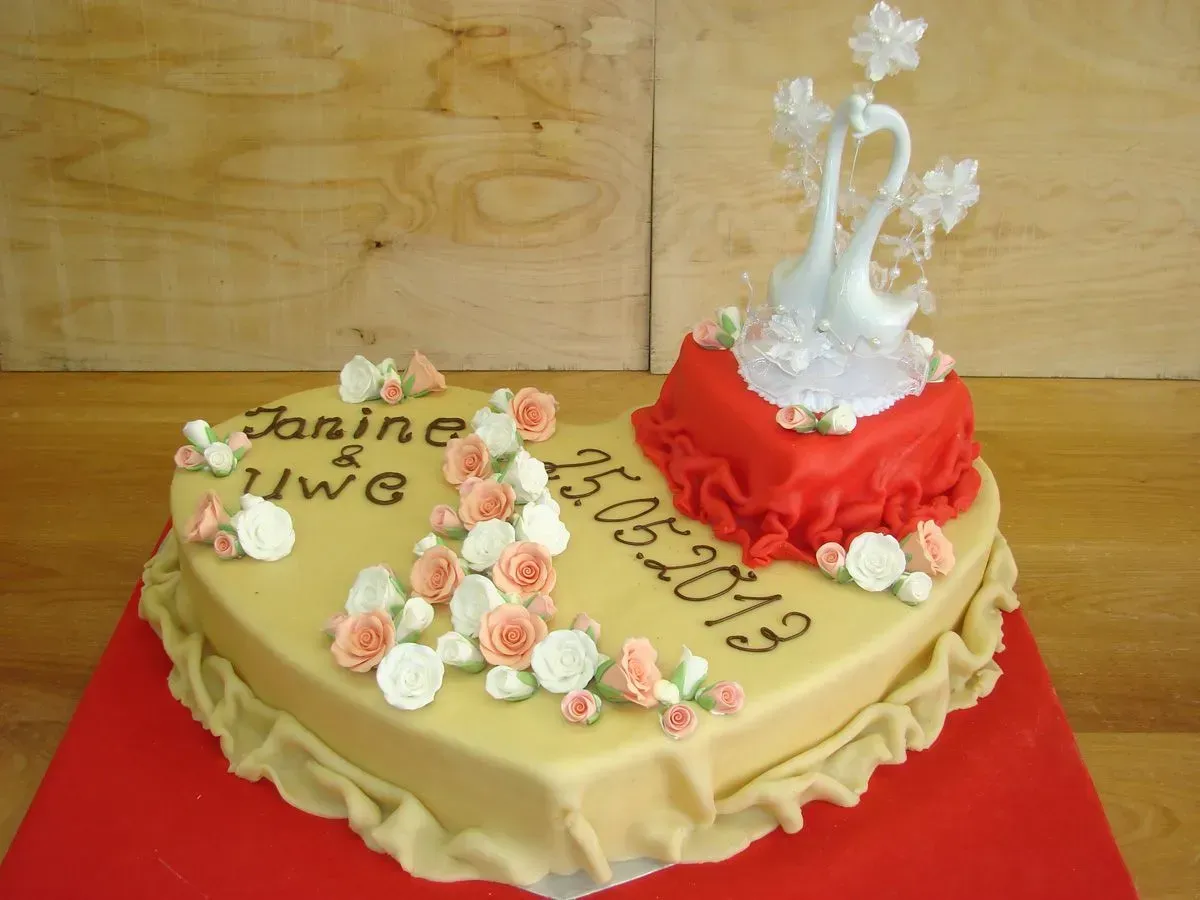 Herzförmige, beige und rote Festtagstorte mit weißem Blumendekor und der Aufschrift „Janice & Vic 10-5-2019“
