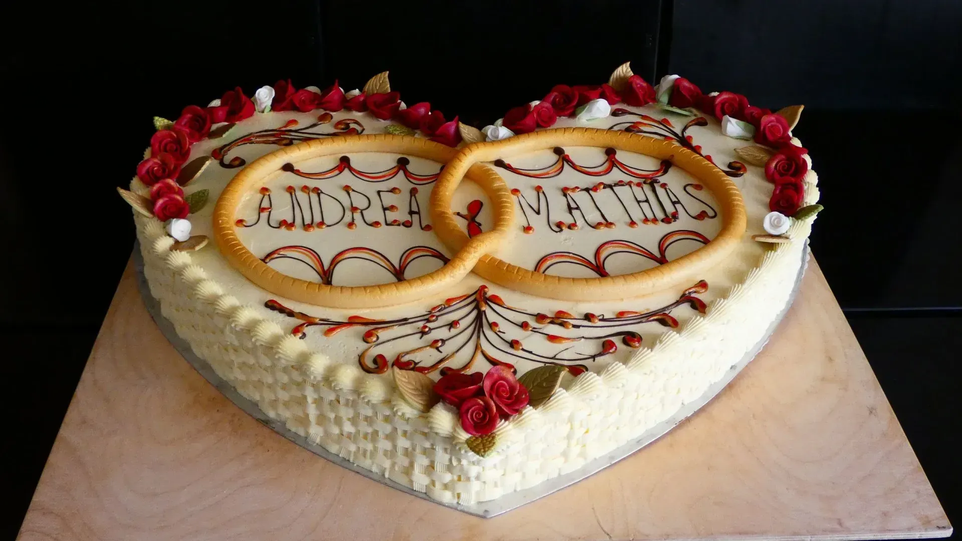 Herzförmiger, verzierter Kuchen mit weißem Zuckerguss, roten Rosen und der Aufschrift „Andrew & Maria“