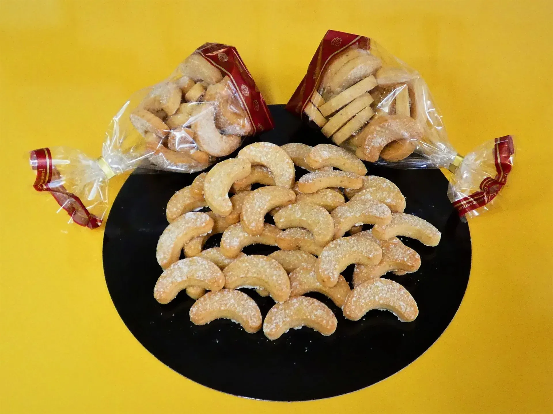 Verschiedene halbmondförmige Kekse auf einem schwarzen Teller, dahinter zwei durchsichtige Snacktüten auf gelbem Hintergrund