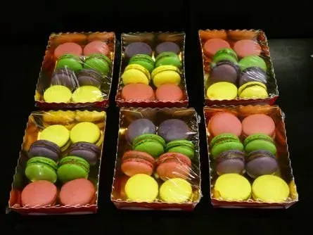 Sechs offene Schachteln mit bunten Macarons, gitterförmig auf einer dunklen Oberfläche angeordnet.