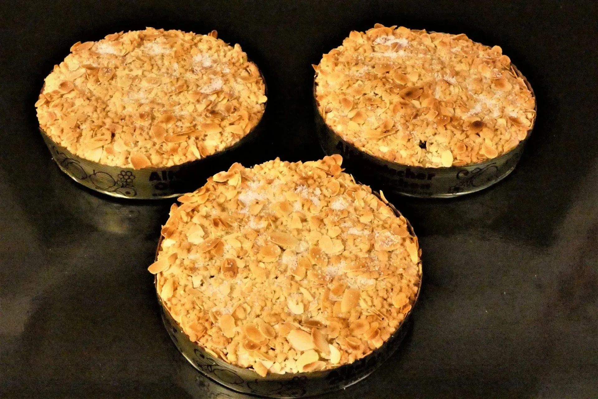 Drei gebackene, mit Streuseln bedeckte Dessertkugeln in dunklen Backformen auf einer schwarzen Oberfläche