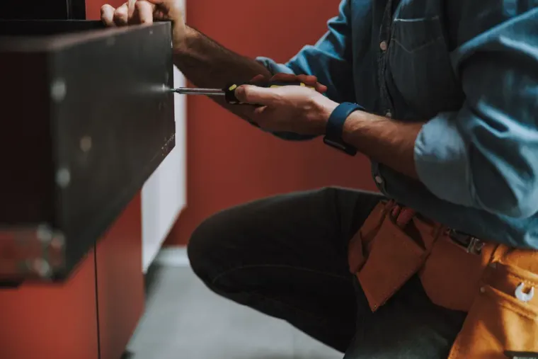 Una persona con camisa azul está en cuclillas, usando un destornillador para reparar un cajón oscuro.