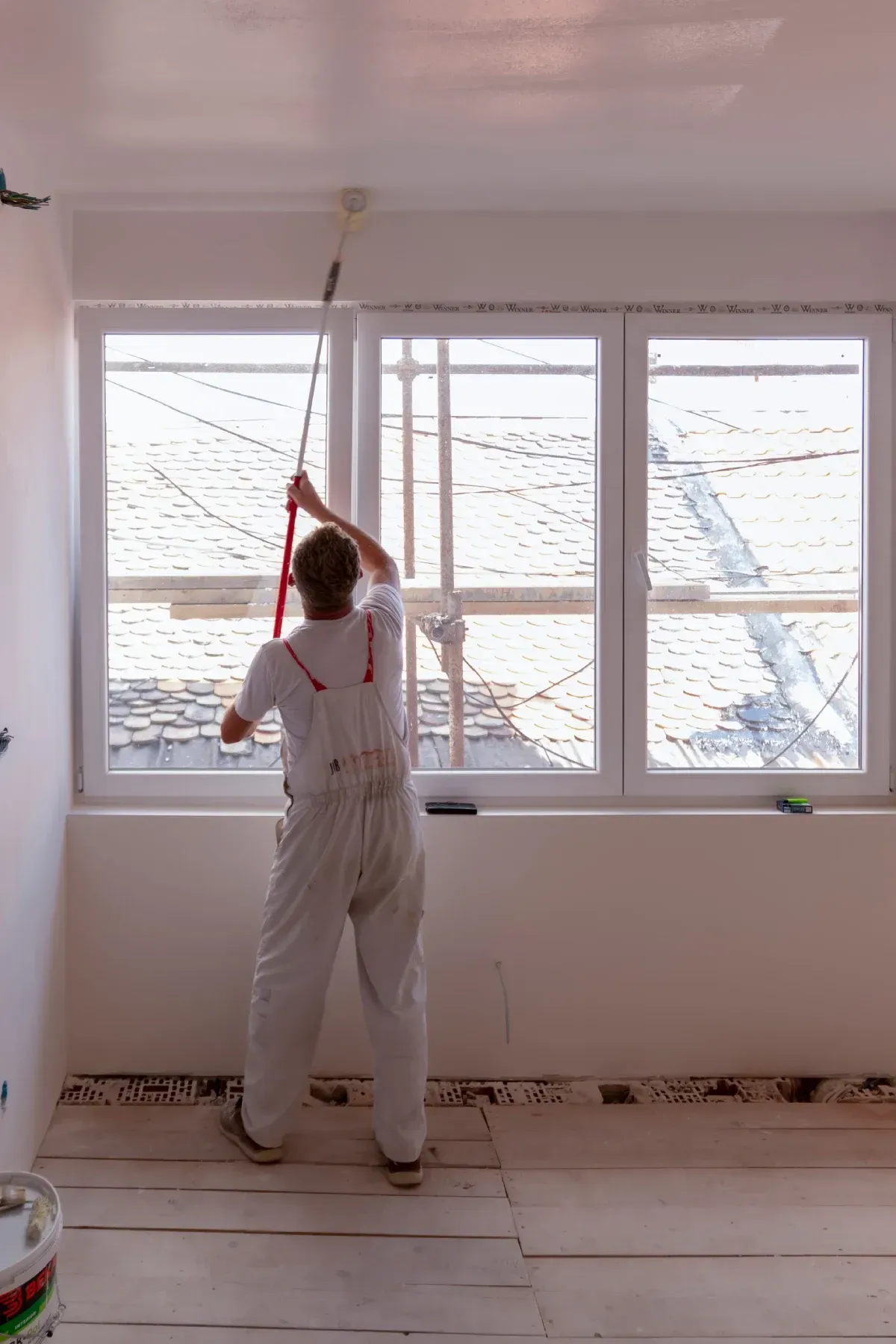 Una persona vestida con un mono blanco pinta un techo con un rodillo, de pie en una habitación con una gran ventana.