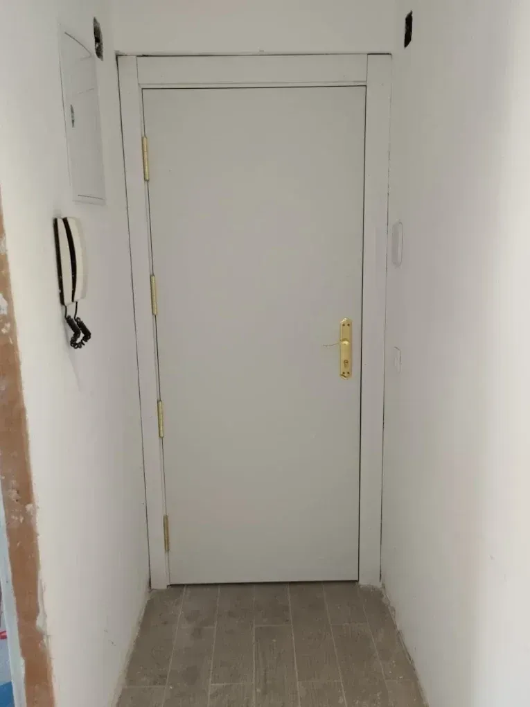 Puerta blanca en un pasillo estrecho con suelo de baldosas, teléfono y cuadro eléctrico en la pared.