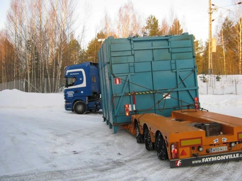 Sininen kuorma-auto vetää sinivihreää ylisuurta kuormaa lumisella tiellä, taustalla paljaita puita
