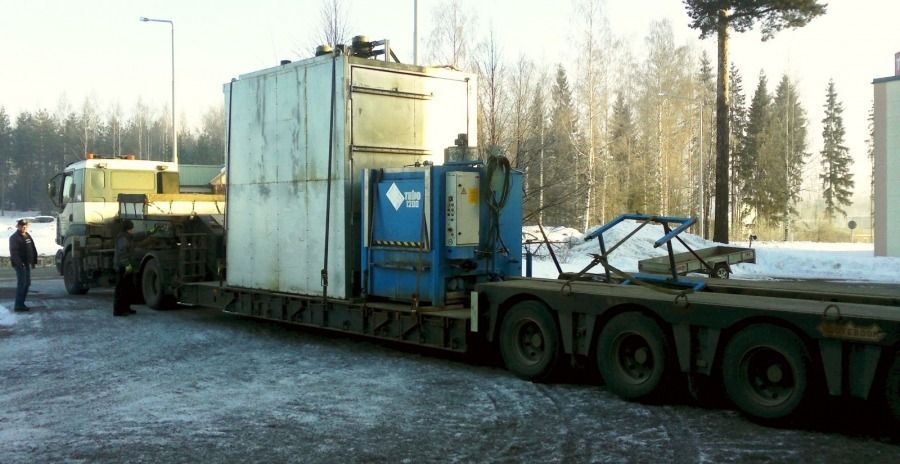 Keltainen kuorma-auto vetää suurta sinistä teollisuussäiliötä lavetilla lumisella tienvarsialueella