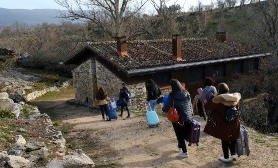 Un grupo de personas con equipaje camina hacia un edificio de piedra con techo de tejas. Al aire libre, por un sendero de tierra.