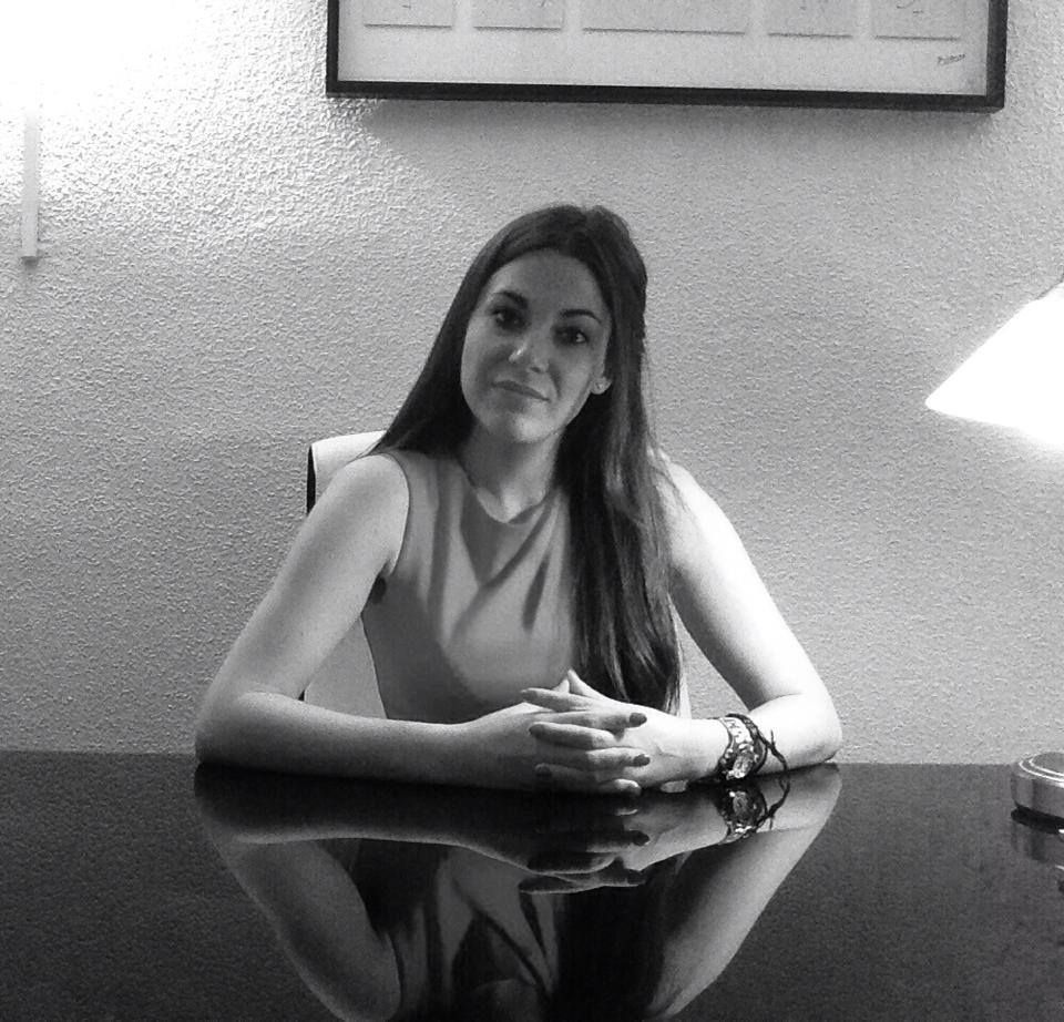 Mujer sentada en un escritorio, con las manos juntas, mirando a la cámara. Oficina, lámpara y obra de arte en la pared.