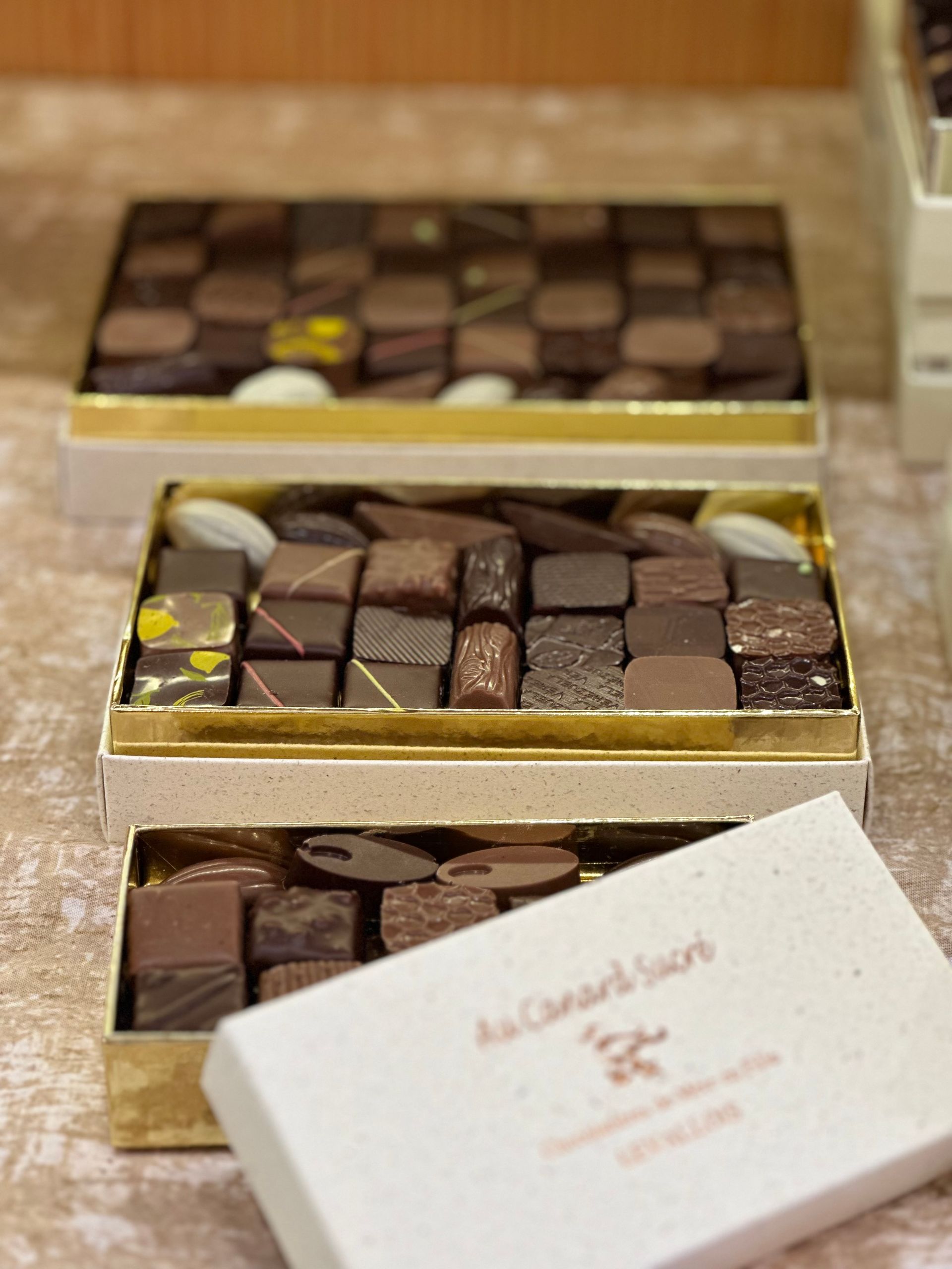 Des boîtes de chocolats dorées ouvertes, contenant des chocolats carrés assortis, sont disposées sur une table.