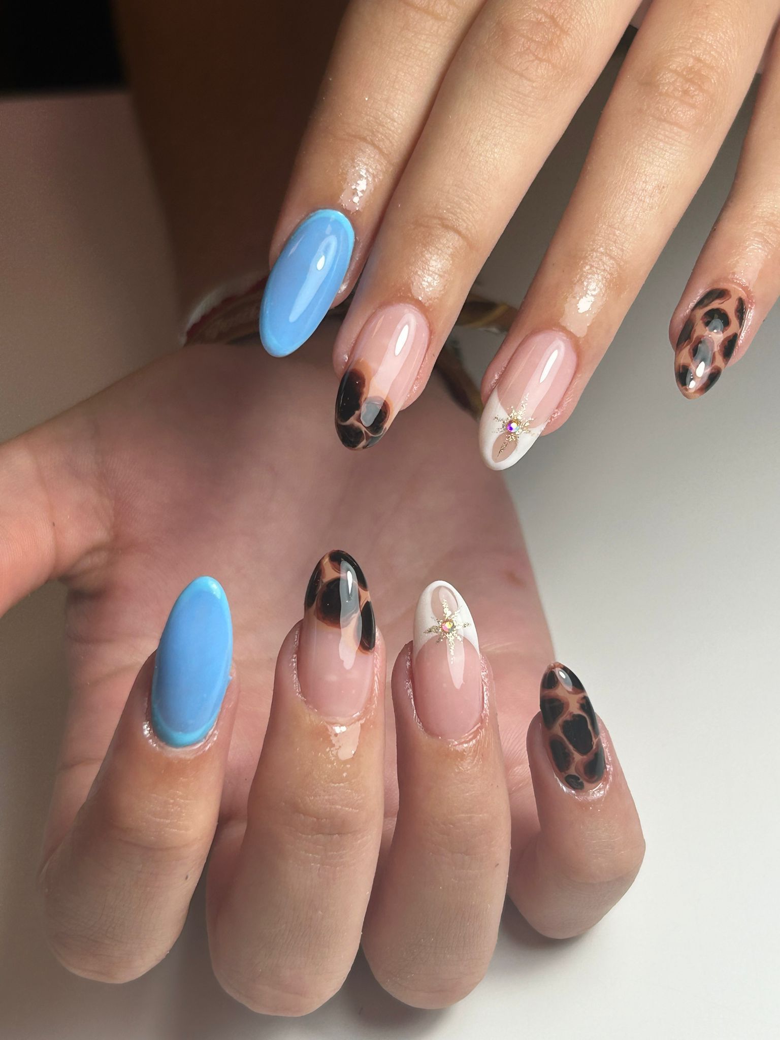 Primer plano de una mano con uñas ovaladas. Los diseños incluyen azul claro, estampado de leopardo y puntas blancas con una gema.