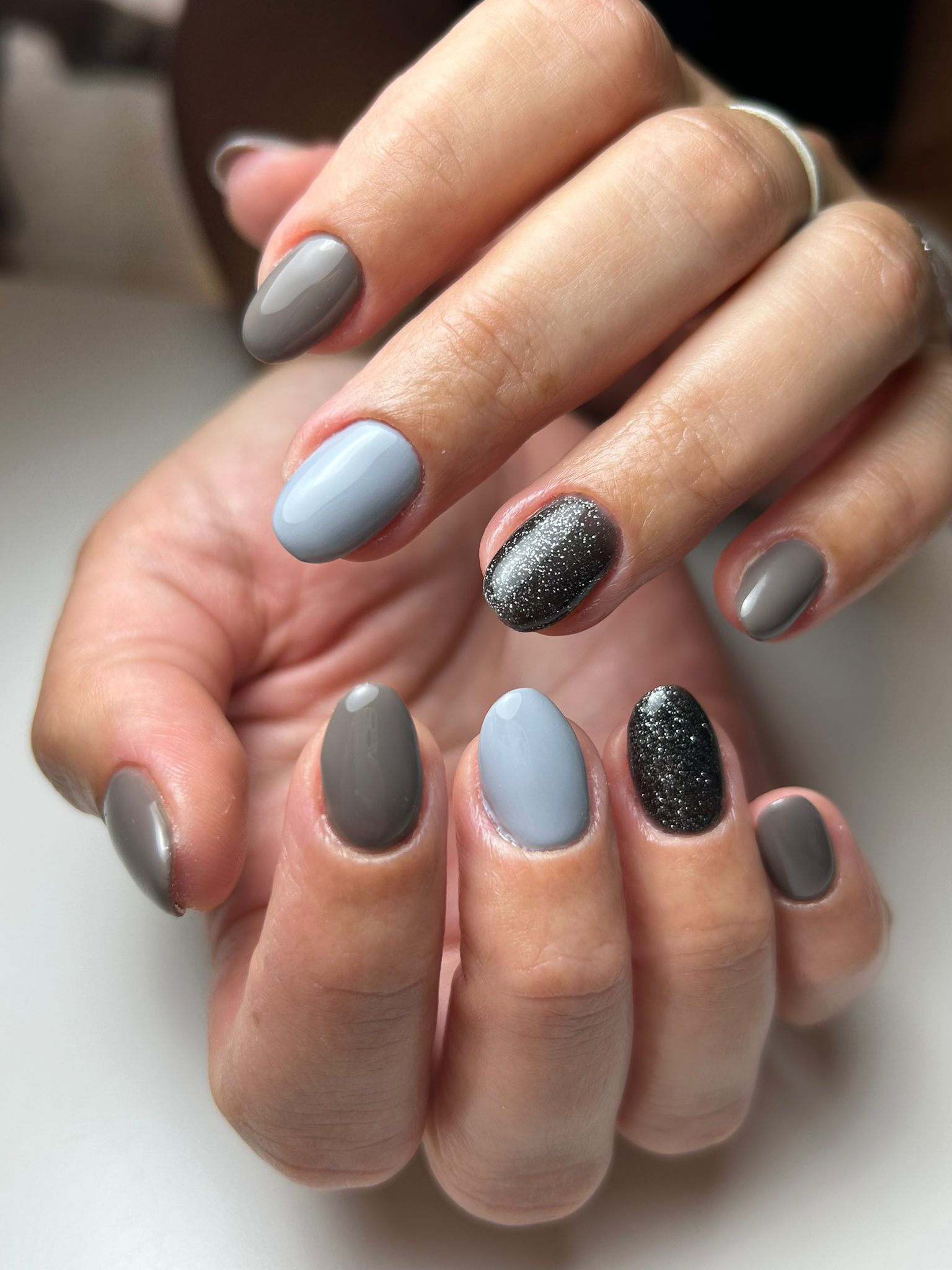 Manos con uñas de forma ovalada, pintadas de gris, azul claro y negro brillante, sobre una superficie blanca.