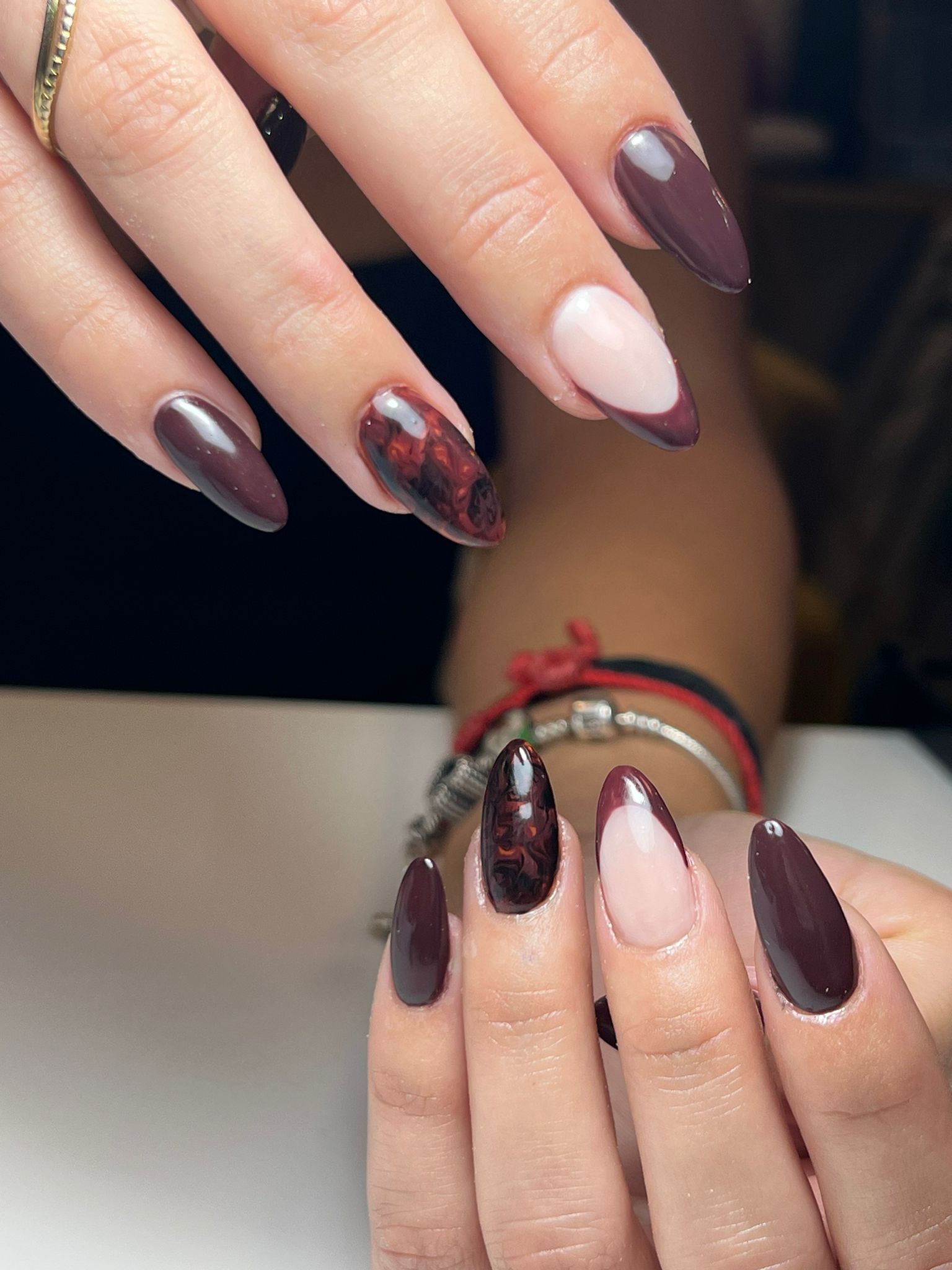 Manos con uñas almendradas pintadas de granate y un detalle detallado. Una uña tiene una punta francesa y la otra un diseño rojizo.