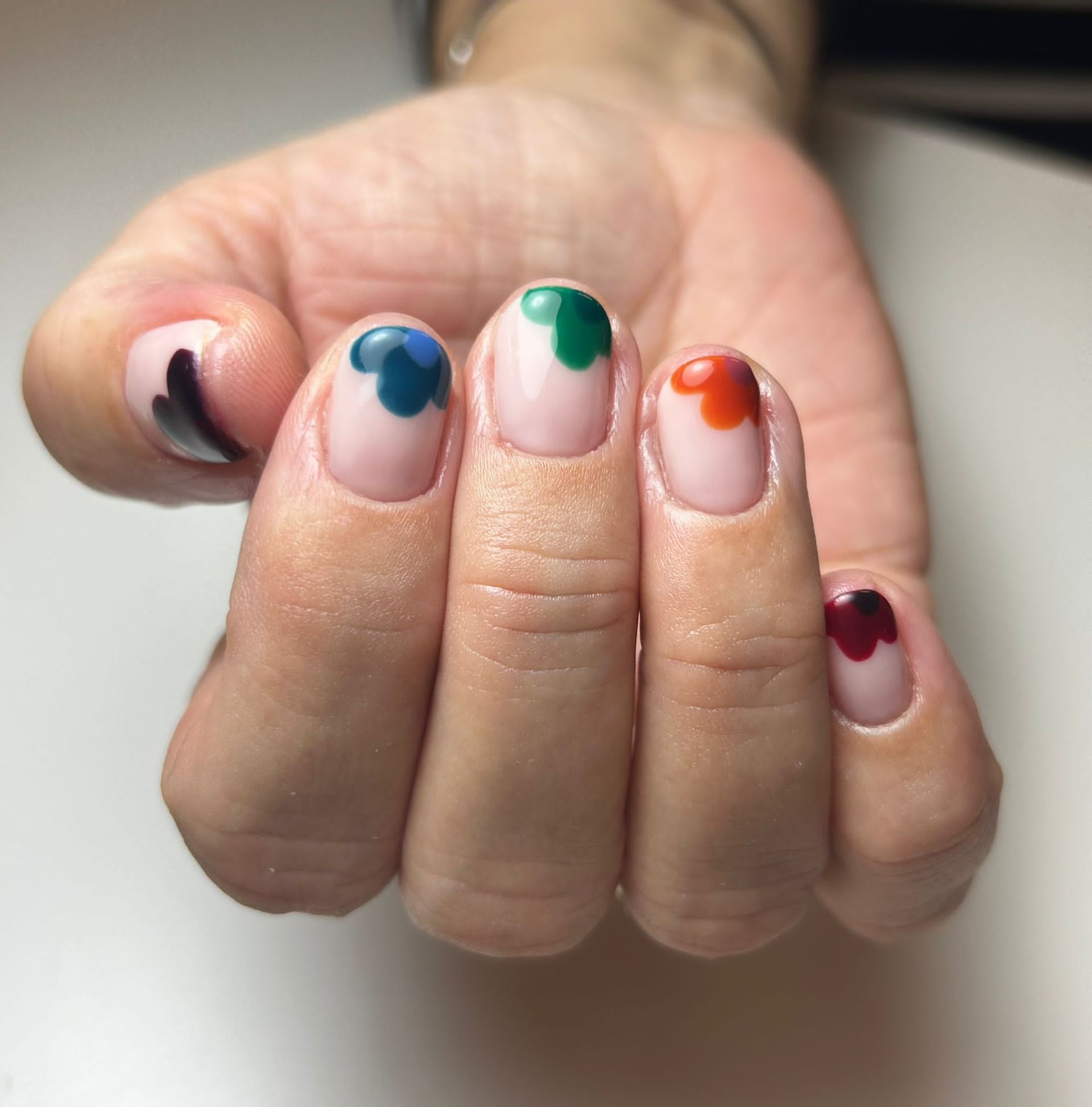 Mano con uñas cortas, cada una con punta redondeada de diferente color: negro, azul, verde, naranja y rojo.