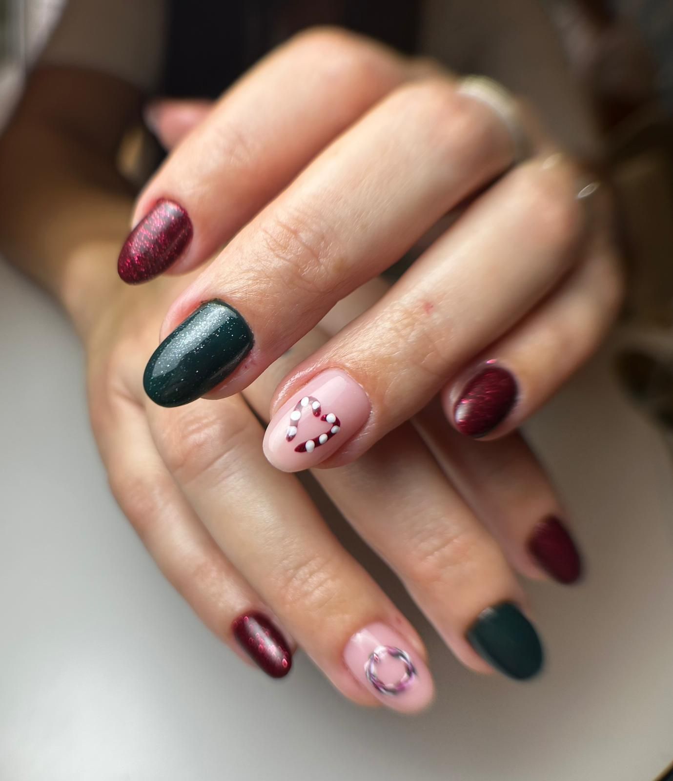 Manos con esmalte de uñas burdeos, verde oscuro y rosa. Dos uñas rosas lucen diseños de bastones de caramelo blancos.
