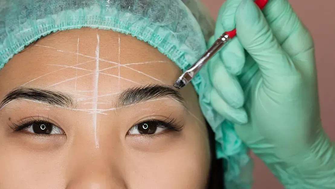 Primer plano de una persona con líneas dibujadas en la frente, a quien alguien que usa guantes le está dando forma a las cejas.