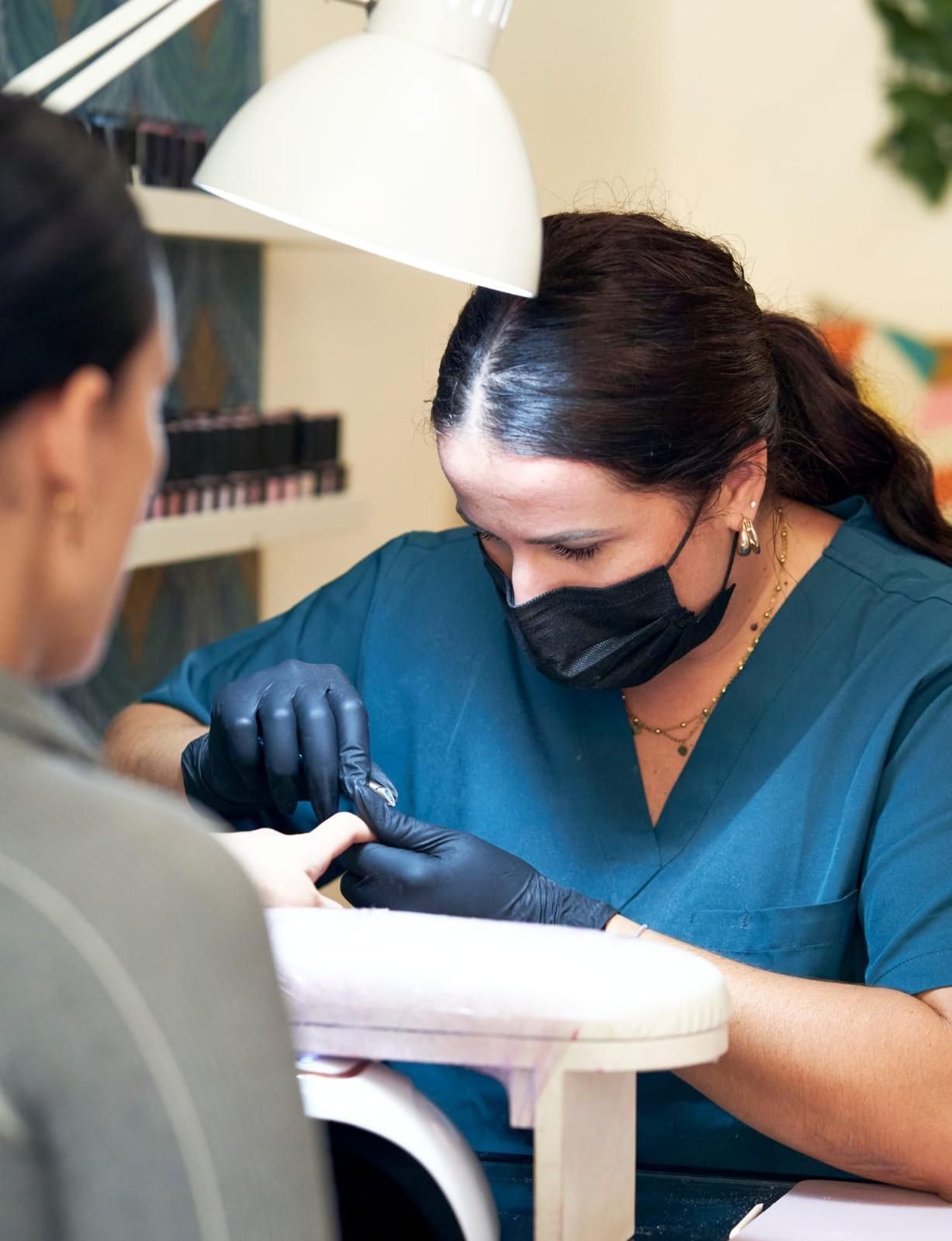 Persona con mascarilla haciendo la manicura a una clienta, usando una luz y trabajando en un salón.