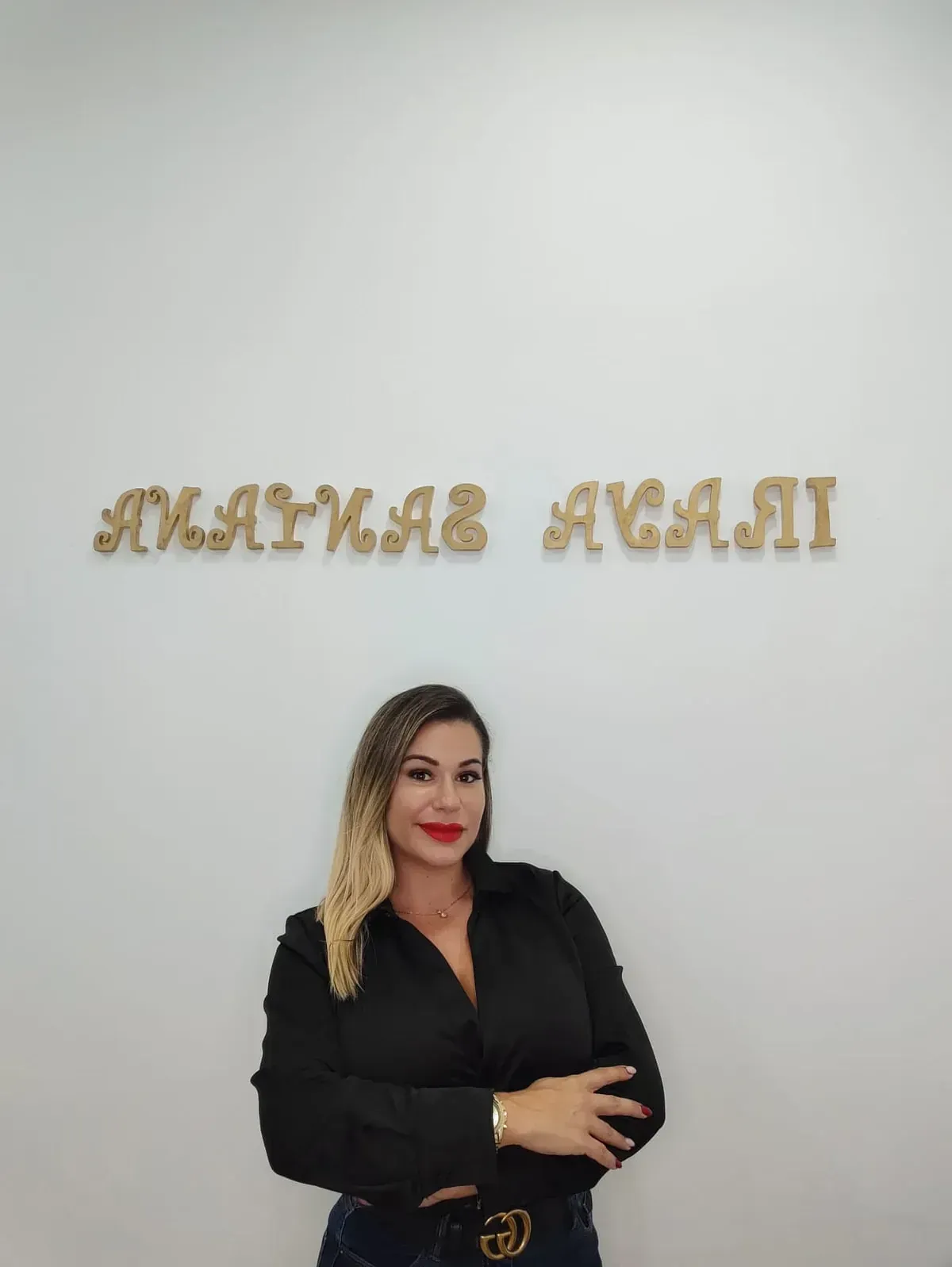 Mujer con camisa negra, posando frente a una pared blanca con letras doradas que dicen 