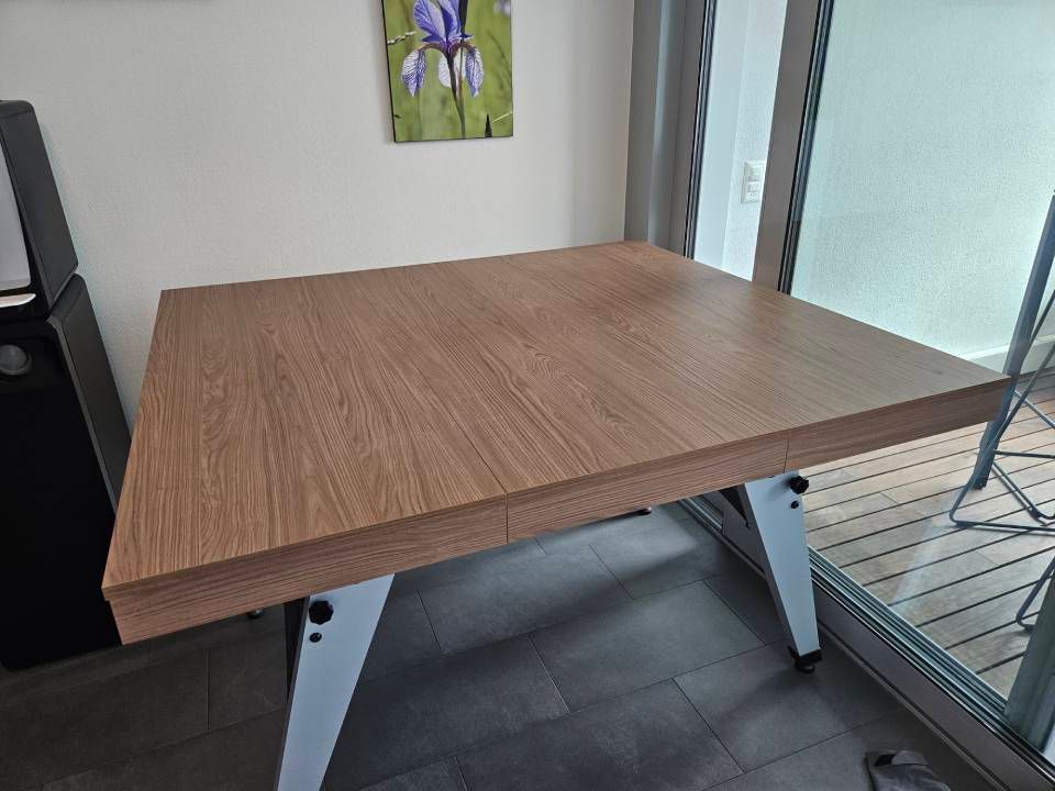 In einem hellen Zimmer in der Nähe einer Balkontür steht ein Holztisch mit dicker Kante und modernen weissen, kantigen Beinen.