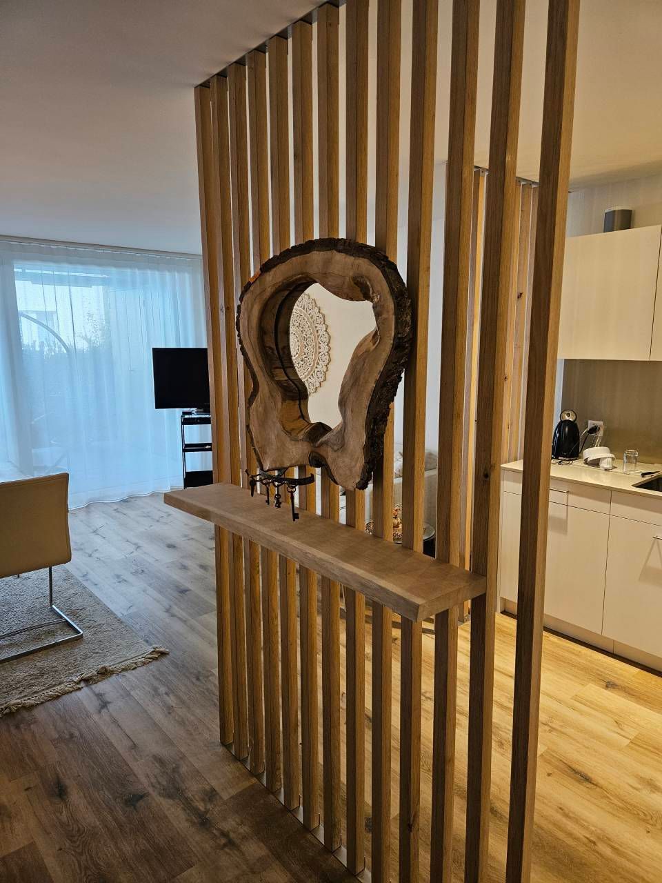 In einem modernen, offenen Wohn- und Küchenbereich steht ein Raumteiler aus Holzlatten mit einem Spiegel mit Baumkante und einem kleinen Regal.