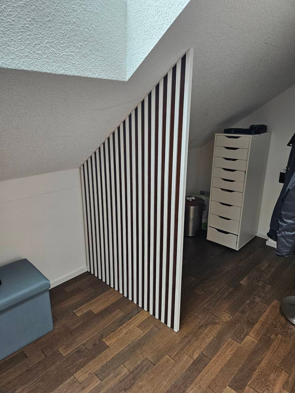 Ein schräges Dachzimmer mit einer dekorativen Raumteilerwand aus Holzlatten, Holzbodenoptik und einem weissen Schubladenschrank.