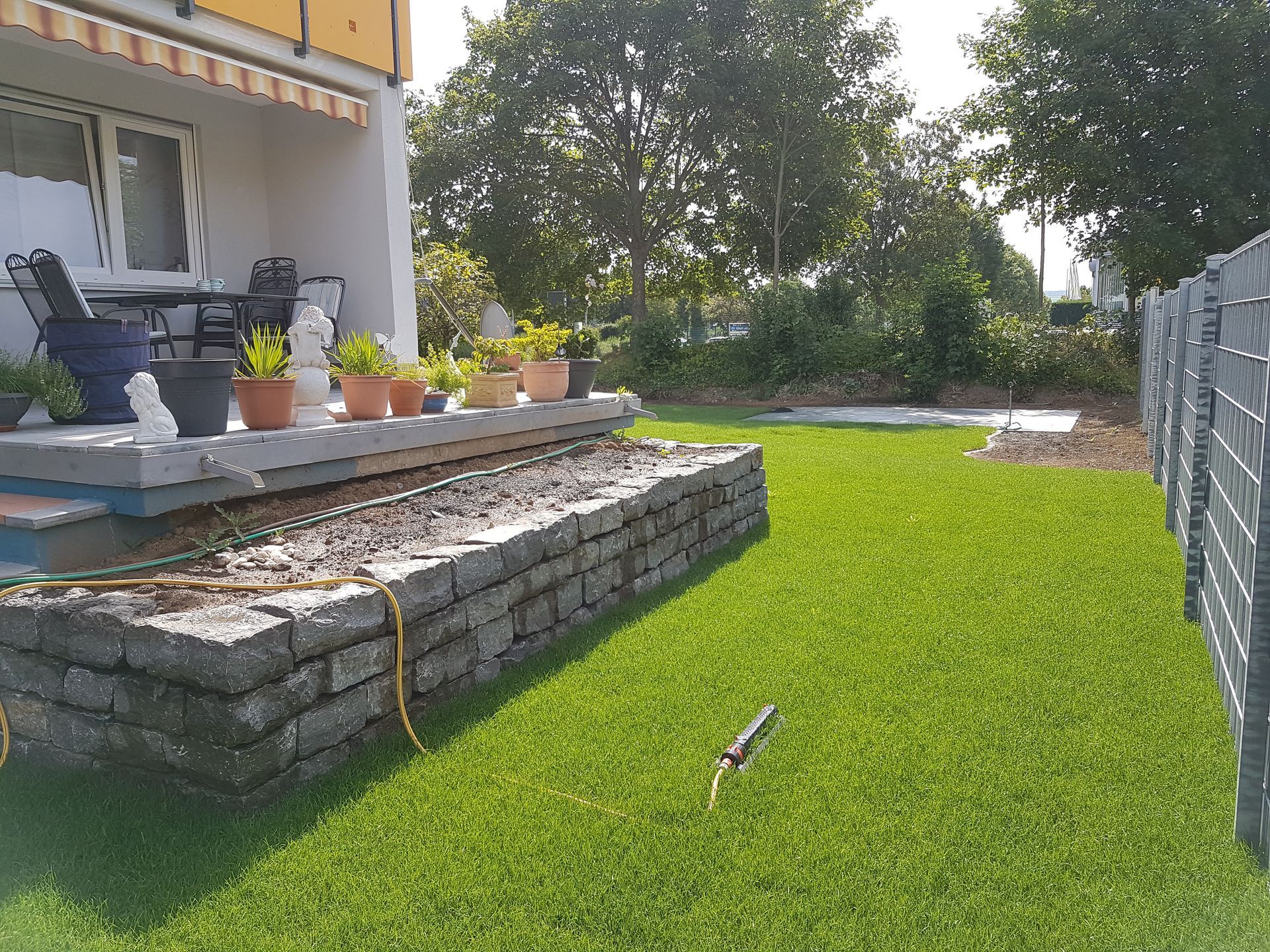 Rasen- und Terrassenbereich mit Topfpflanzen, Steinmauer und Metallzaun.