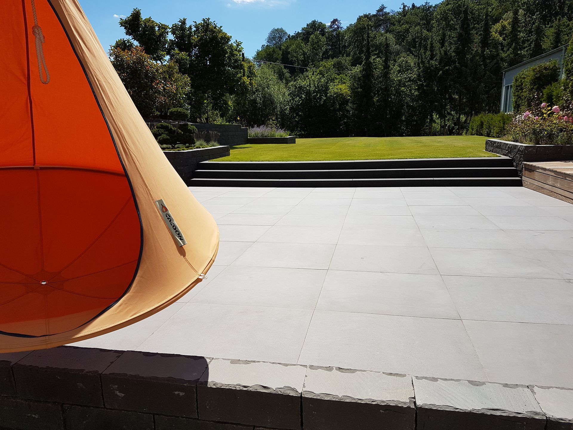 Oranger Hängesessel auf grauer Terrasse mit Blick auf grünes Gras und Bäume an einem sonnigen Tag.